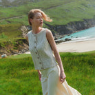 A woman stands in a grassy coastal landscape wearing a sleeveless textured light beige vest with large buttons down the front and cream wide-leg trousers. The background features green hills, a sandy beach, and turquoise water.