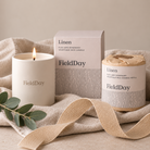 A FieldDay linen scented candle set including a lit white soy wax candle, its matching outer box, and a refill jar wrapped in kraft paper, styled on a beige linen cloth with a ribbon and eucalyptus leaves.